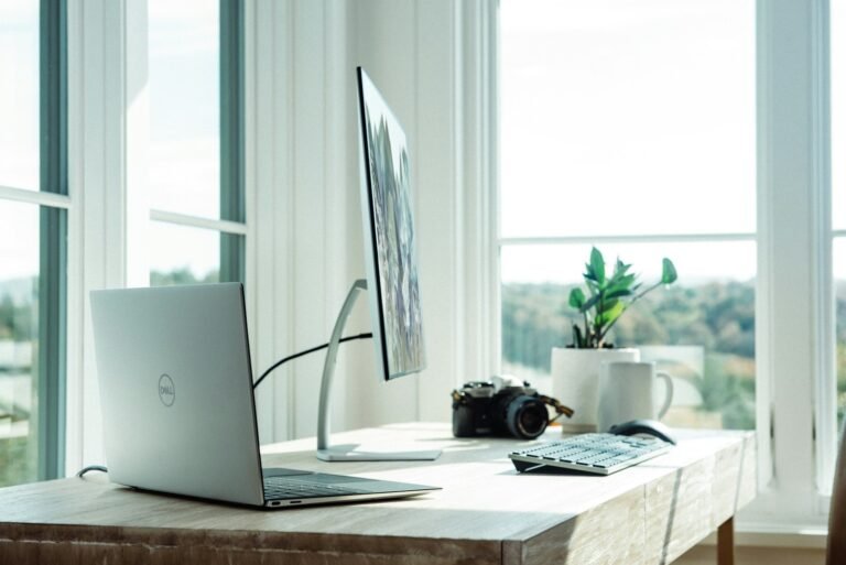 A pale wooden desk in front of tall windows, with a laptop, a monitor on a slim stand, a wireless keyboard, a camera, a mug and a small plant