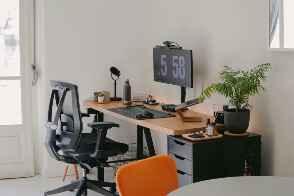 A real home office setup: a black mesh office chair with adjustable armrests at a wooden desk, a monitor raised on a stand, a desk microphone and keyboard, a potted plant on a grey drawer unit, and daylight coming in from a window and door to the left