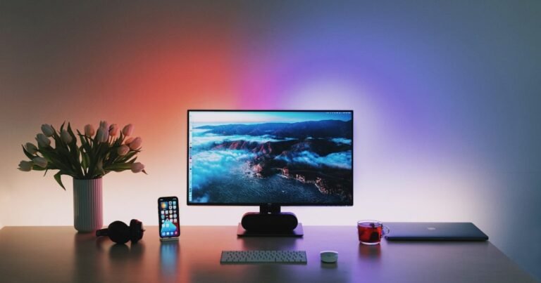 A single monitor on a dark desk with a keyboard, mouse, phone and a closed laptop, with coloured bias lighting washing the wall behind