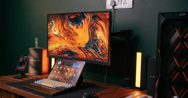 A gaming laptop open on a desk in front of a larger external monitor, with a mechanical keyboard, a mouse and a light bar