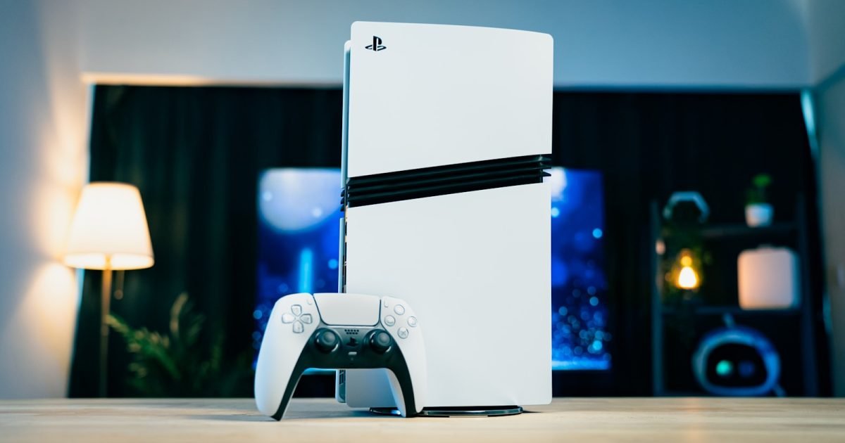 A white PlayStation 5 standing upright on a wooden desk with its controller in front and a lamp glowing to one side